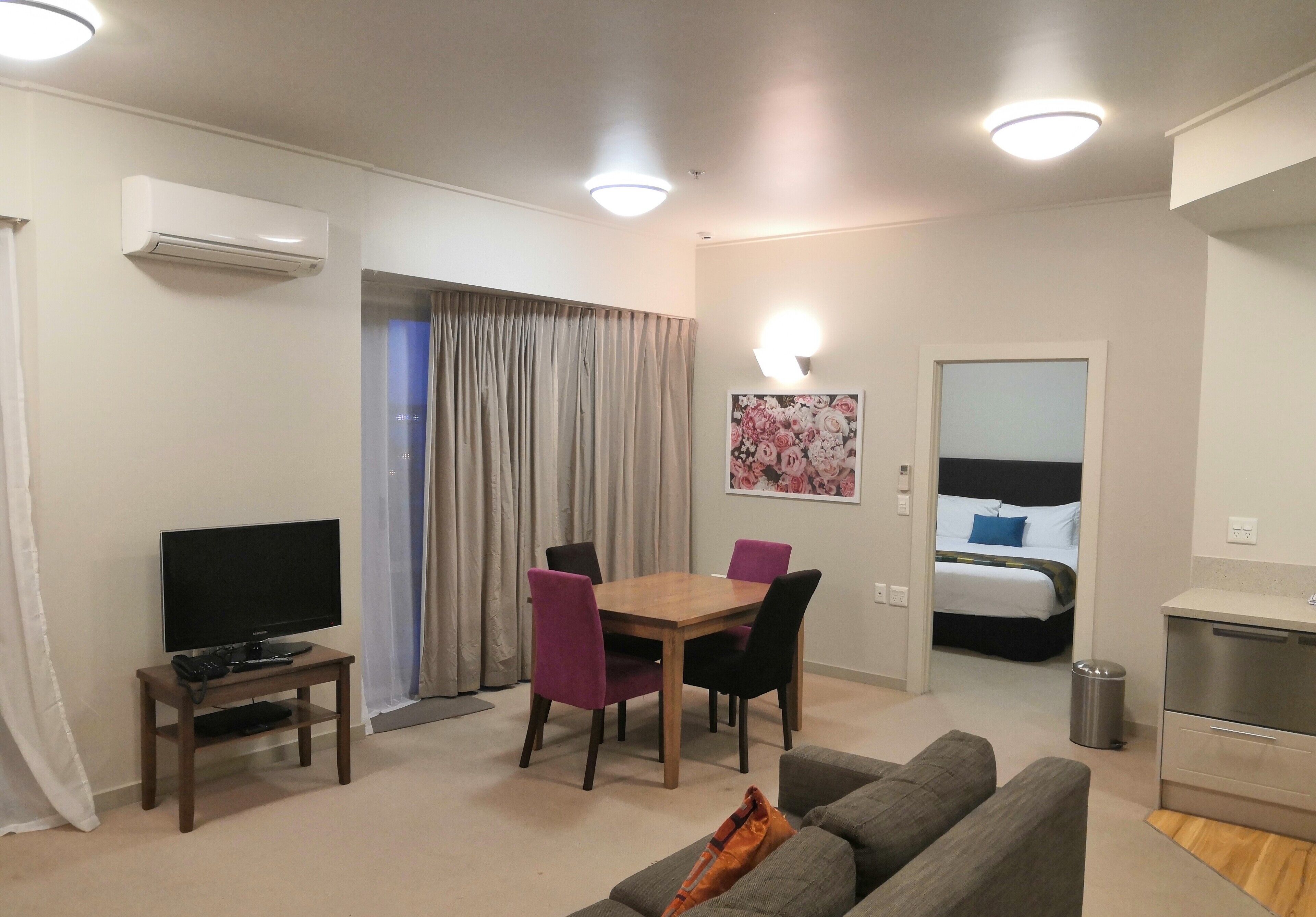 A modern living room with a flat-screen TV, a wooden dining table with four chairs, a grey sofa, a small kitchen area with a stainless steel refrigerator, and a bedroom with a bed and a nightstand.