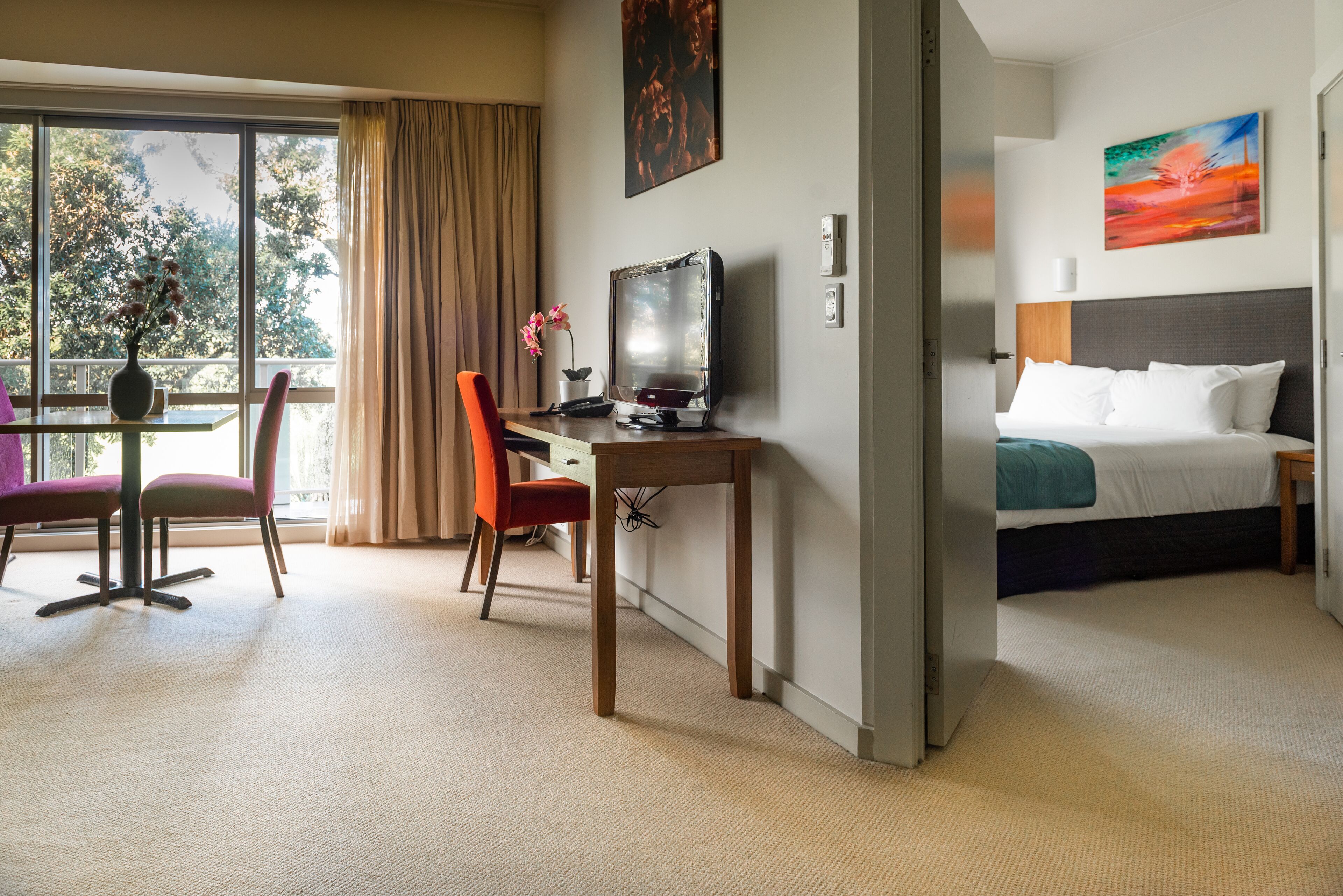 A hotel room with a bed, a desk with a computer, a chair, a window with curtains, and a painting on the wall.