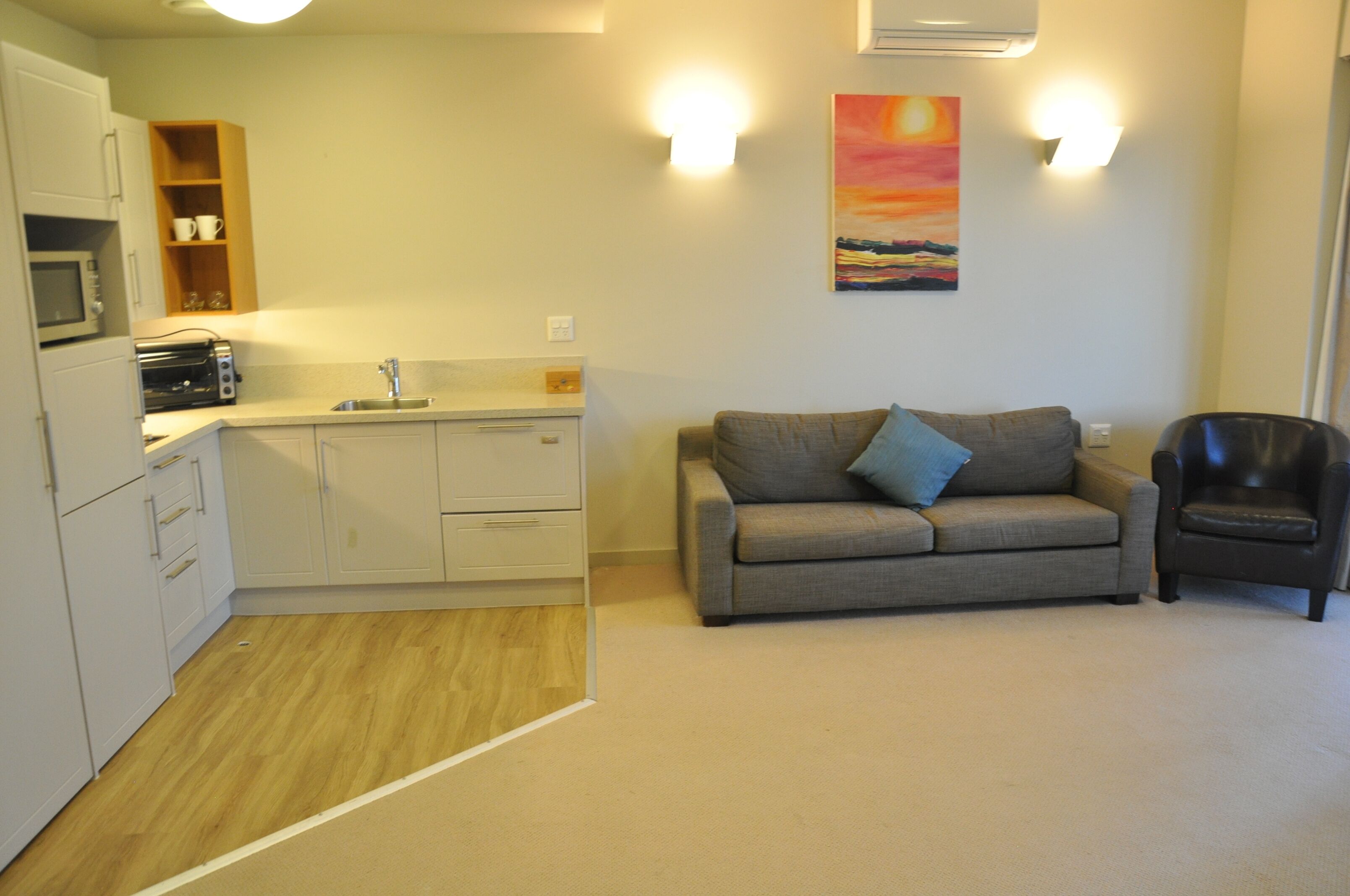 A compact living space with a kitchenette, a sofa, a chair, and a painting on the wall.