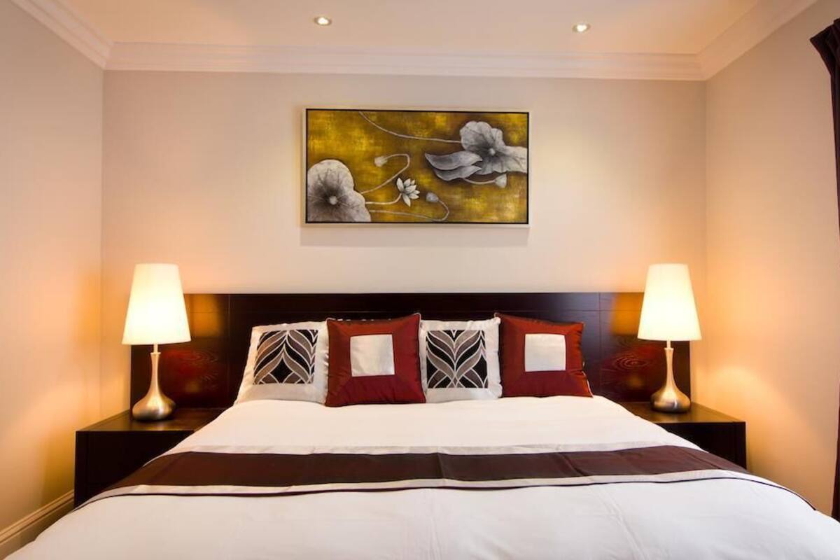A hotel room with a large bed, two bedside tables with lamps, a decorative wall piece, and a window with curtains.