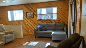 Ein gemütliches Wohnzimmer mit einer Holzwand, einem grauen Ecksofa, einem grauen Sessel und einem kleinen Tisch.