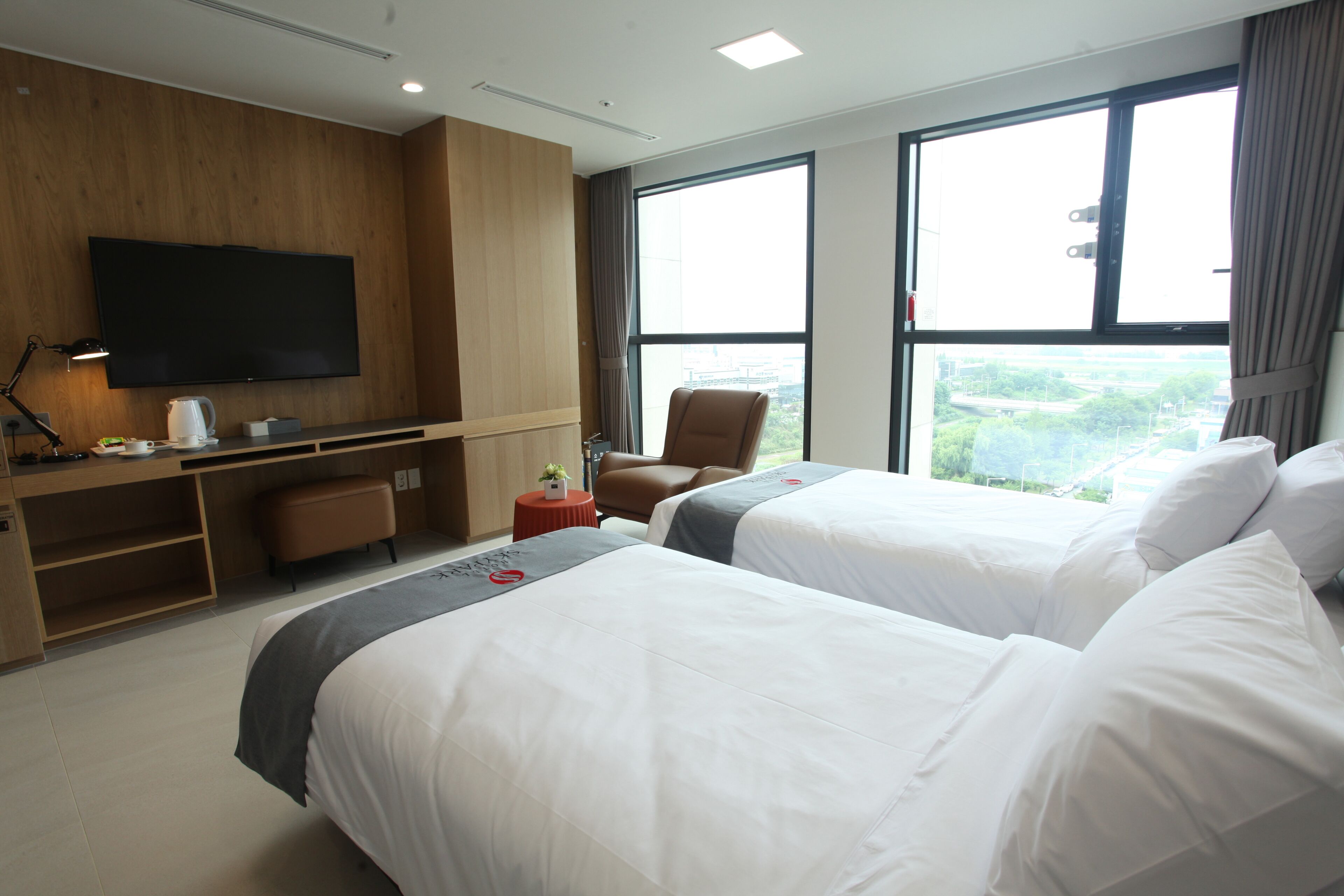 A hotel room with two beds, a TV, a desk, and a view of the city.