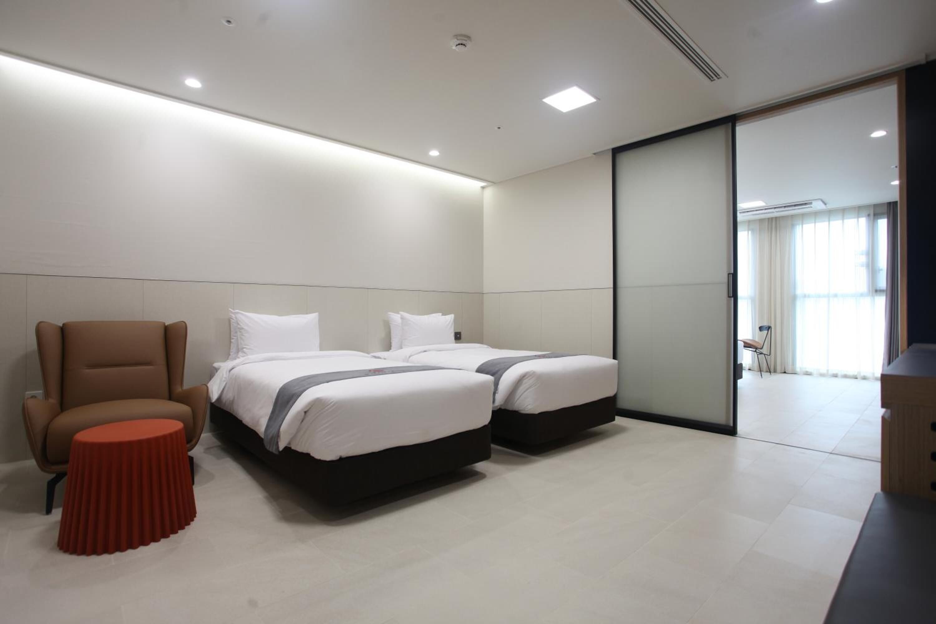 A modern hotel room with two beds, a brown armchair, and a small red table.