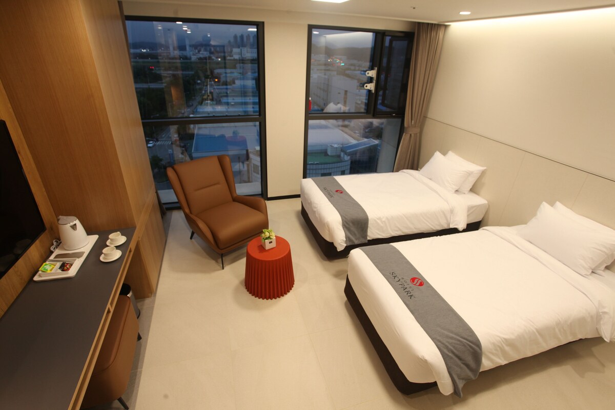 Een hotelkamer met twee bedden, een bureau met koffiezetapparaat, een stoel en een kleine tafel met een plant.