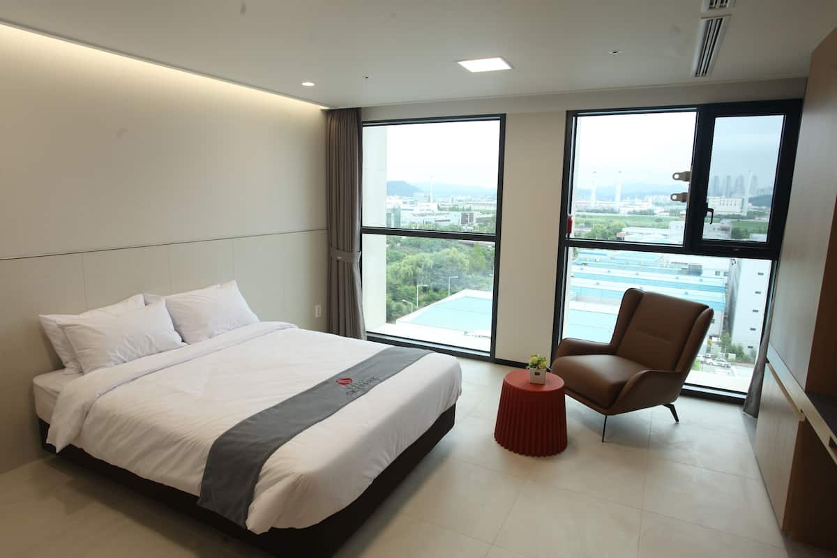 Een moderne hotelkamer met een groot bed, een klein rond tafeltje met een vaas, een stoel en grote ramen met uitzicht over de stad.