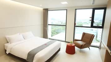 Quarto de hotel com uma cama grande, uma mesinha redonda com um vaso, uma poltrona de couro e janelas amplas com vista para a cidade.