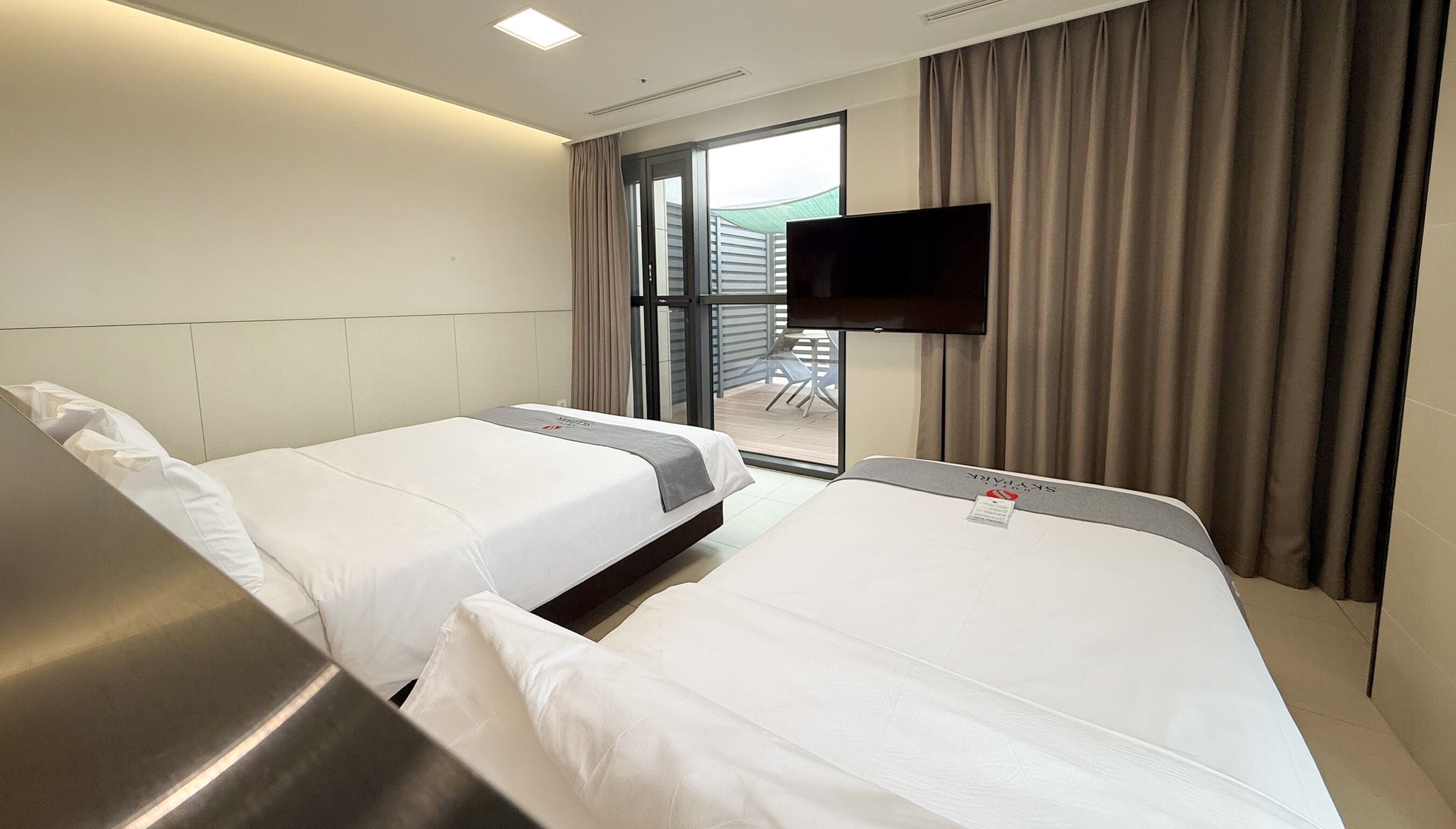 Une chambre d’hôtel avec deux lits, un téléviseur à écran plat fixé au mur et une porte-fenêtre coulissante donnant sur un espace extérieur.