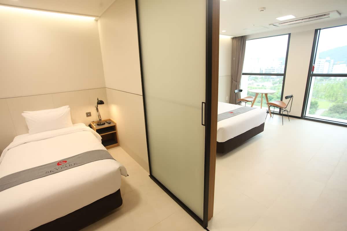 Een hotelkamer met twee bedden, een schuifdeur, een kleine tafel met stoelen en uitzicht op de stad door grote ramen.