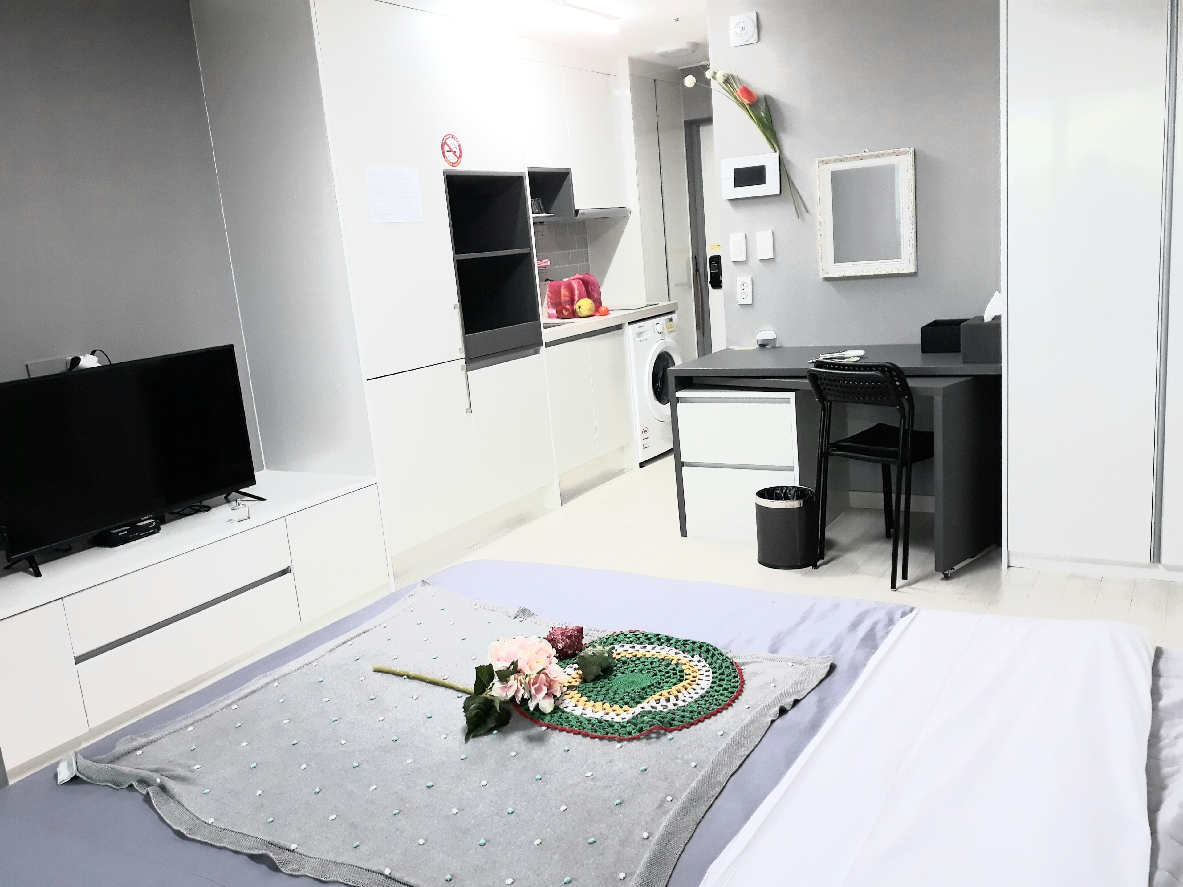 Una habitación de hotel moderna con una televisión de pantalla plana, un escritorio, una silla, un cubo de basura pequeño y un adorno de crochet en la cama.
