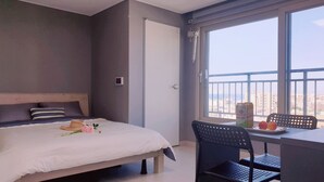 Una habitación de hotel con cama, escritorio con silla, ventana con vista a edificios y una puerta.