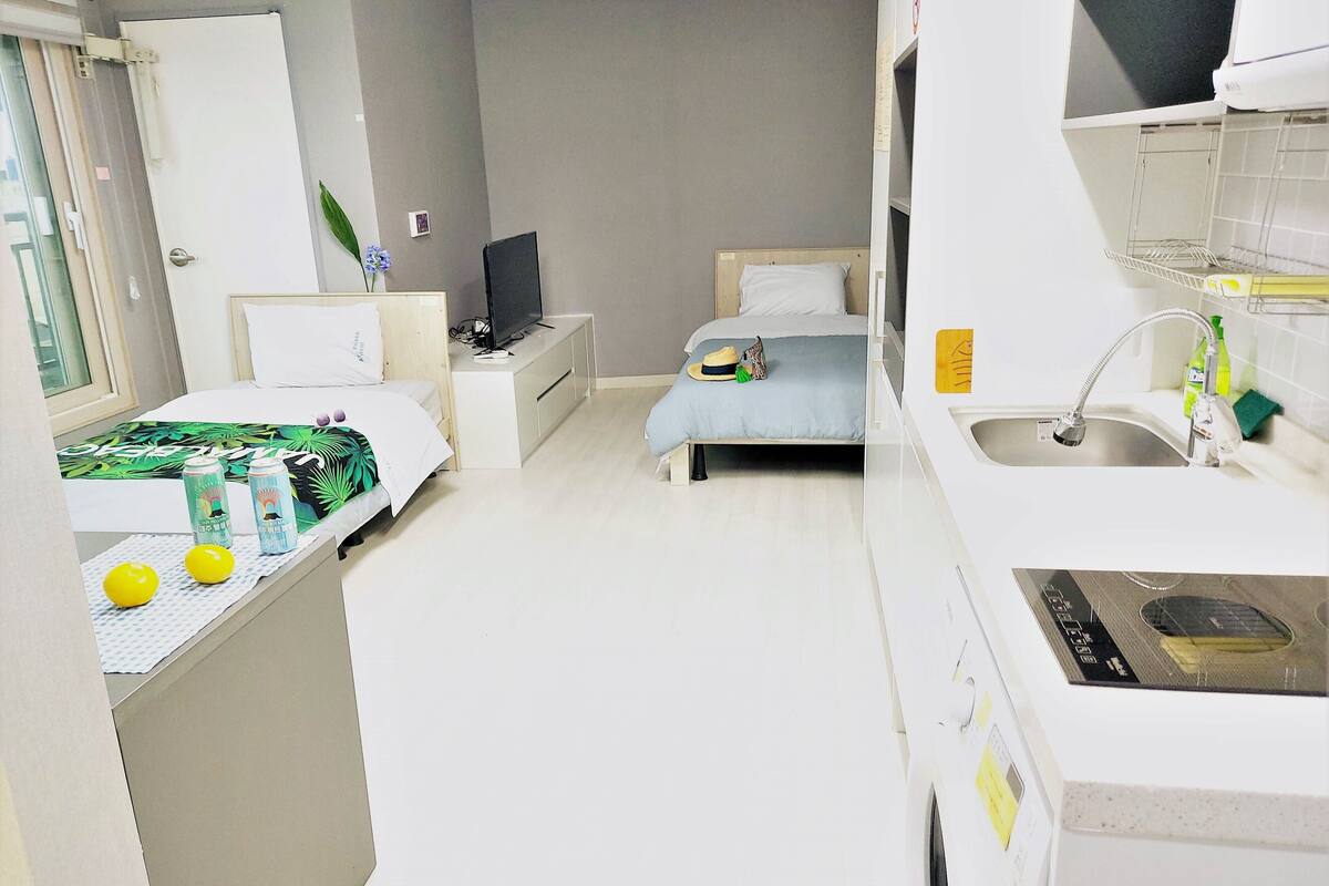A compact hotel room with a bed, a small desk with a TV, a kitchenette, and a bathroom.