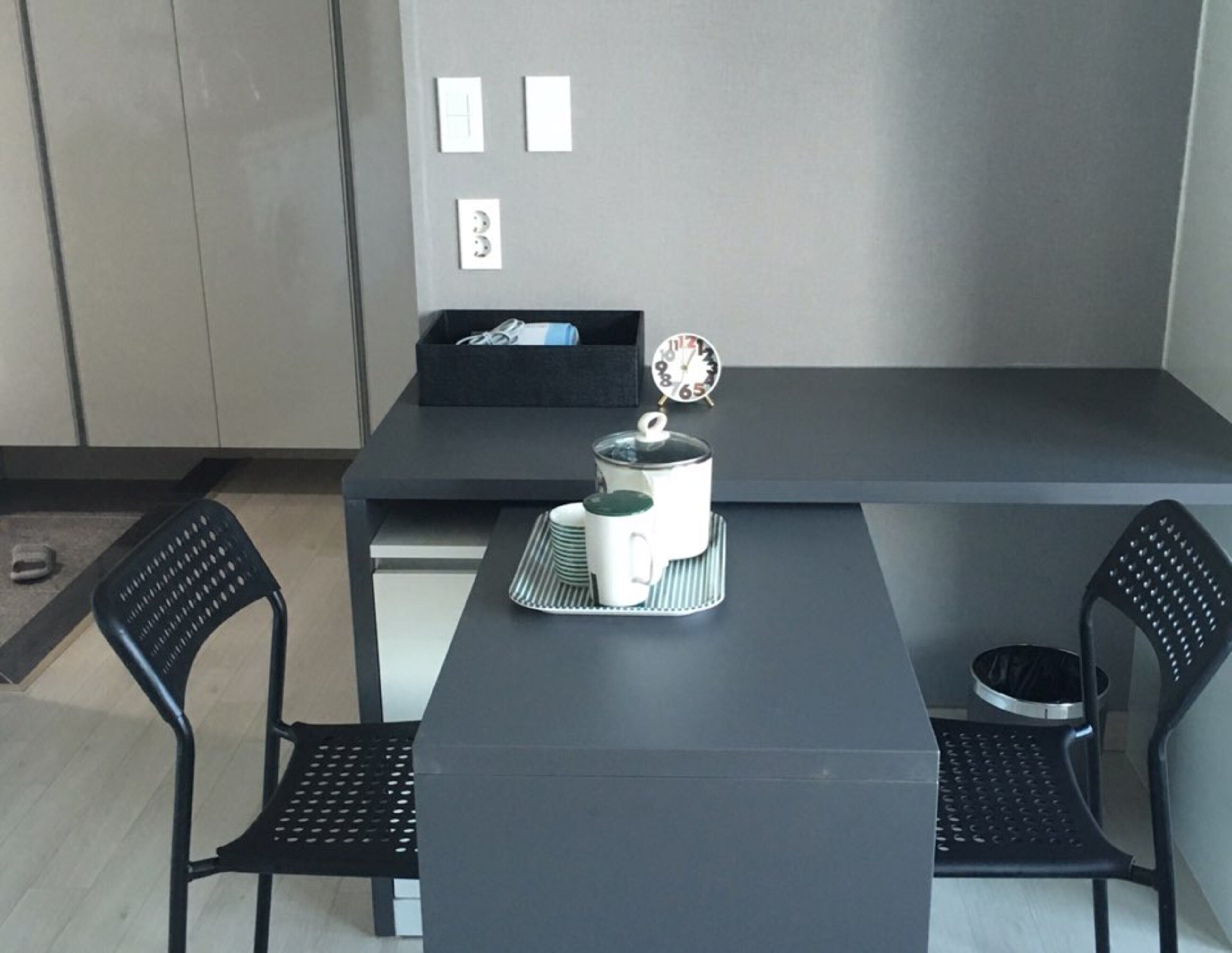 A modern, minimalist room with a dark grey table and two black chairs. A white kettle and a small clock are placed on the table.