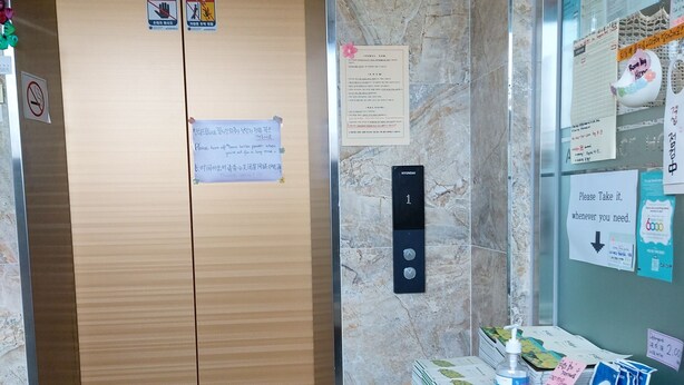 Un hall d’hôtel avec un ascenseur en bois, un paillasson, un panneau d’affichage où sont accrochées diverses annonces, et une petite table sur laquelle sont disposés des articles de toilette et une boîte de mouchoirs.