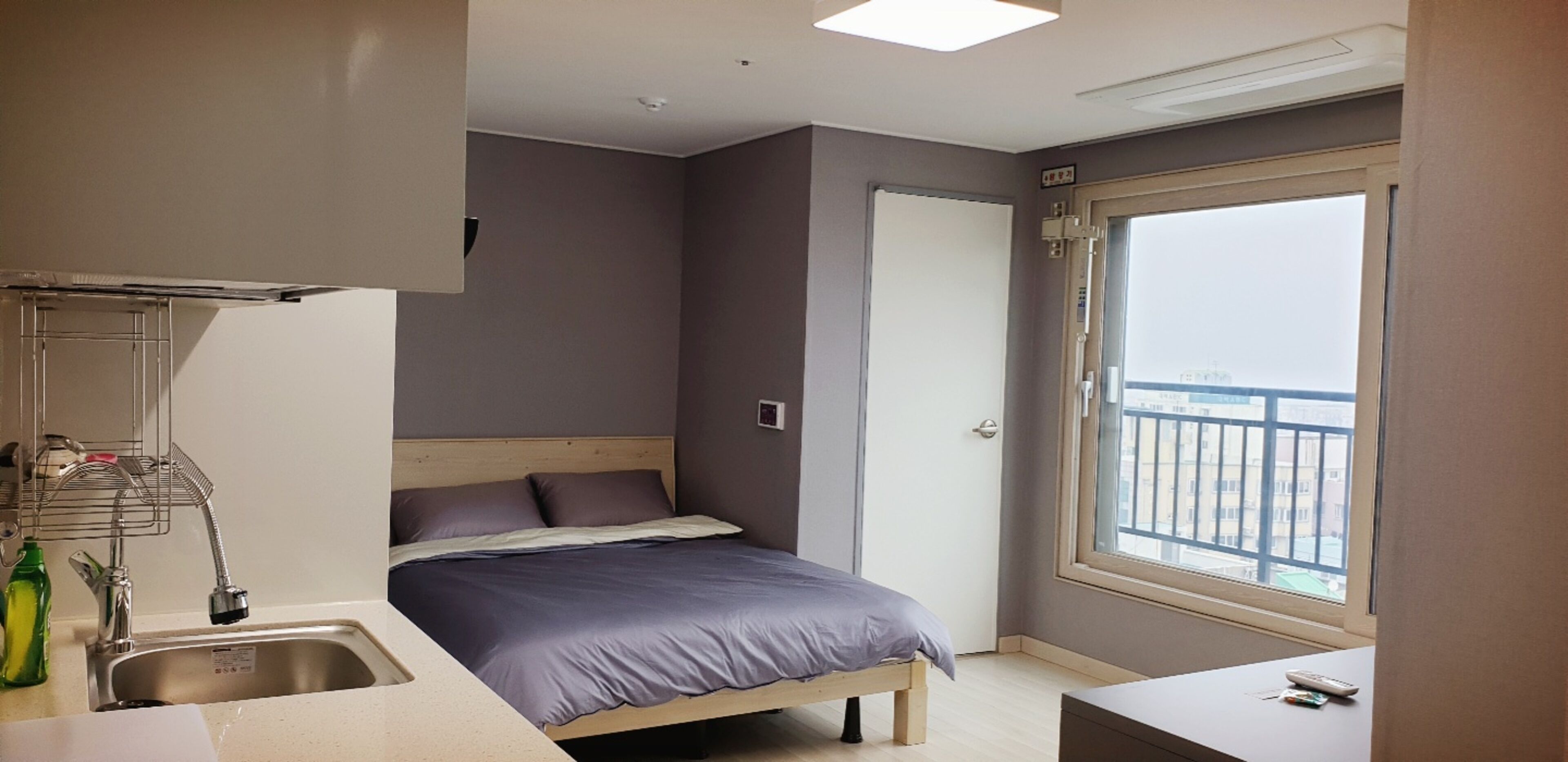 A compact bedroom with a bed, a small sink, and a window with a view of buildings.
