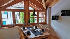 A dining area with a wooden table set for four, surrounded by chairs. Large windows with wooden frames provide natural light and a view of greenery outside.
