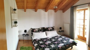A bedroom with a wooden ceiling, a floral bedspread, two bedside tables, a green rug, and a window with curtains.