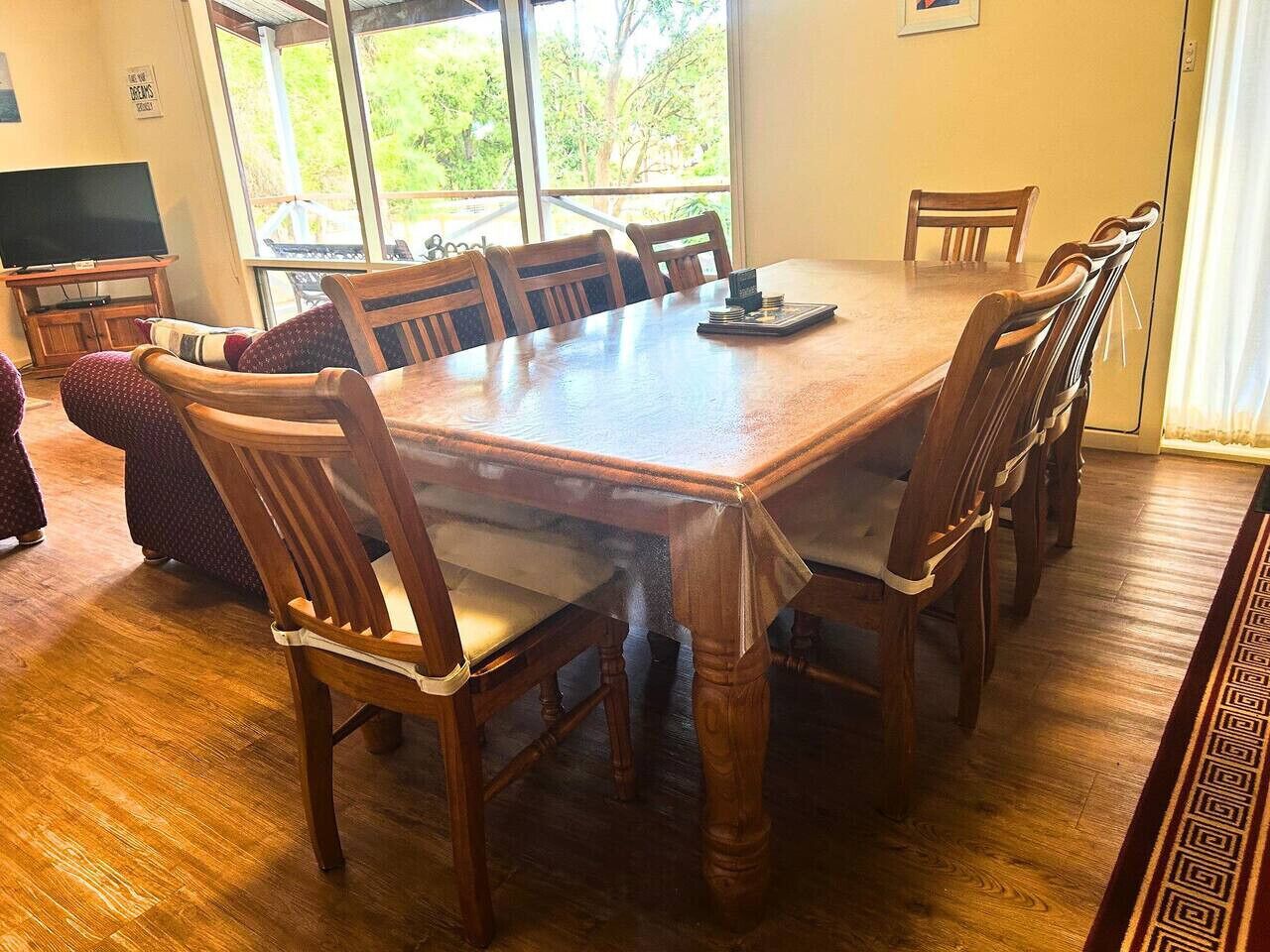 A dining area with a wooden table and chairs, a television, and a view of greenery outside.