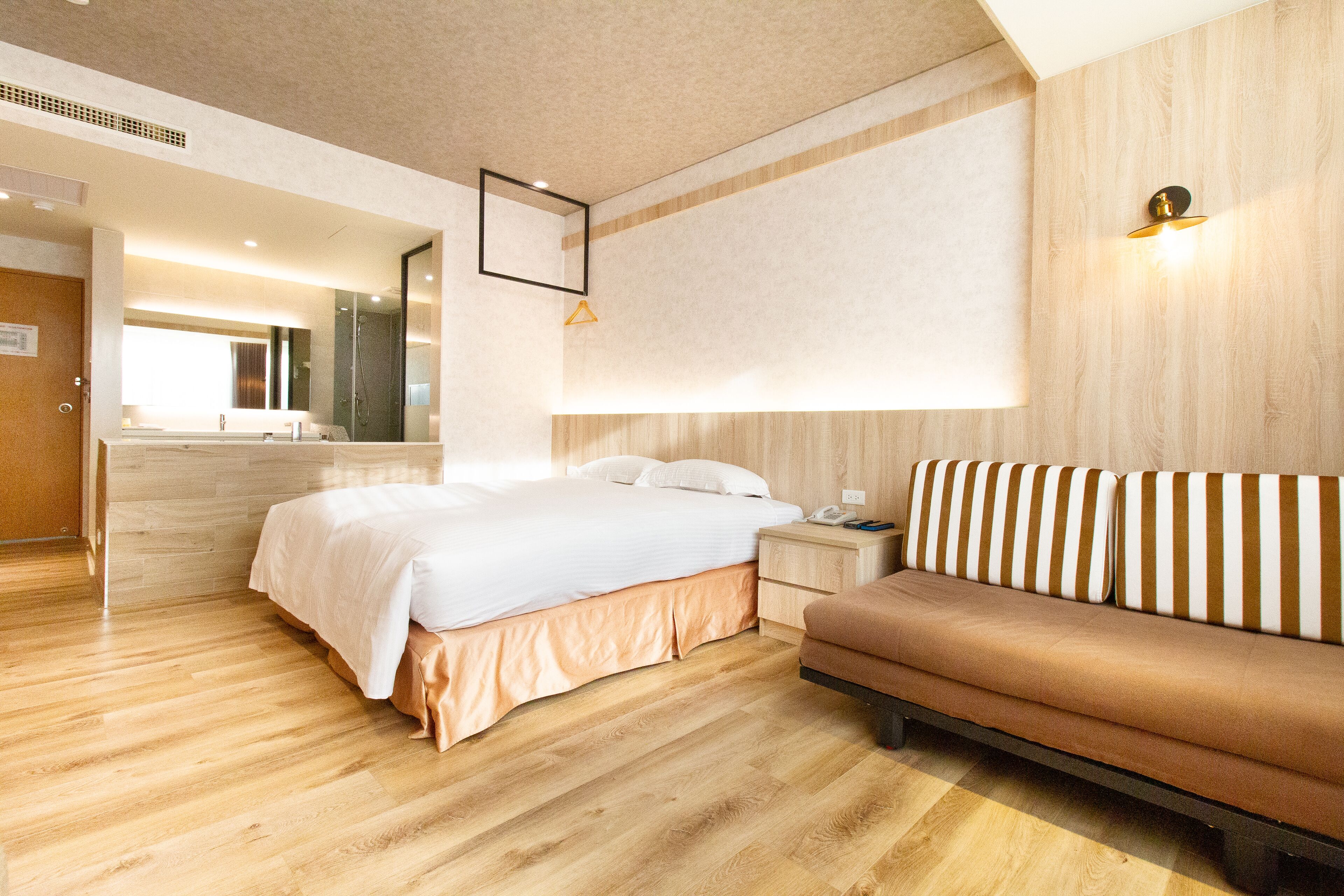 Una habitación de hotel moderna con piso de madera, una cama grande con ropa blanca, un sofá marrón, una mesita de noche con lámpara y un espejo en la pared.