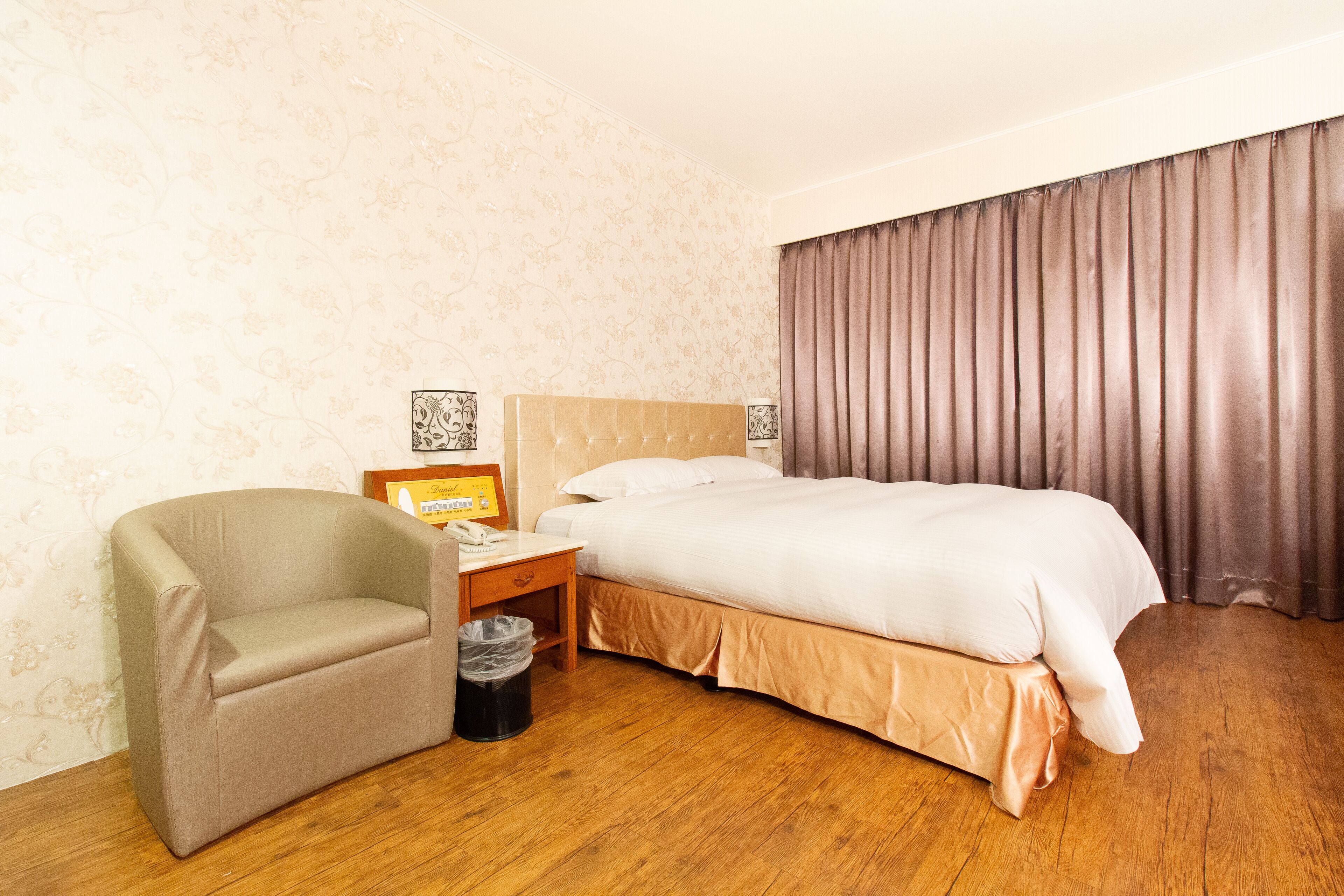 Quarto de hotel com cama, cadeira, mesa de cabeceira e lixeira.