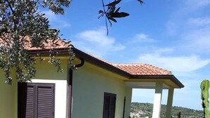 Una casa de dos pisos con balcón, un muro de piedra y una planta de cactus.