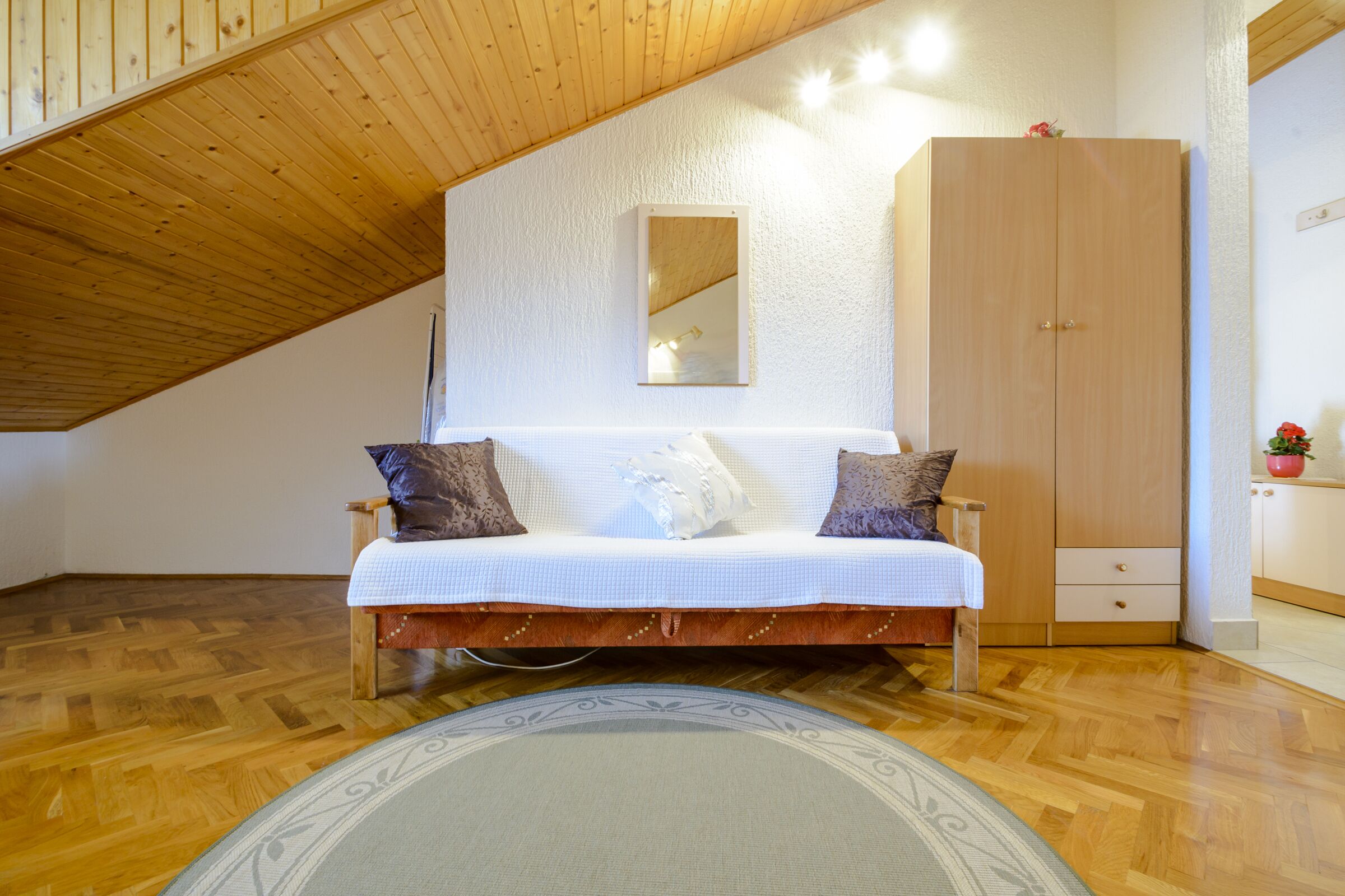 Un dormitorio tipo loft con techo de madera, un sofá blanco con cojines estampados, un espejo grande y un armario.