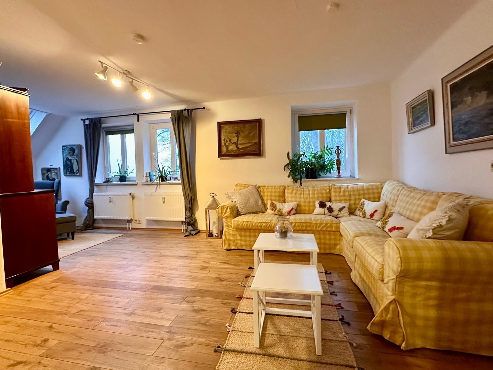 Una sala de estar acogedora con un sofá amarillo a cuadros, una mesa de centro blanca y piso de madera. Se observan ventanas con cortinas, un radiador y una planta en el alféizar.