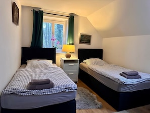 A bedroom with two single beds, a nightstand with a lamp, a window with curtains, and a small rug on the floor. - Wohnung Lotte Petersen - Willi Ohler Haus (Worpswede)