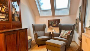 Quarto de sótão aconchegante com claraboia, um armário de madeira, duas poltronas, uma mesinha e um radiador.