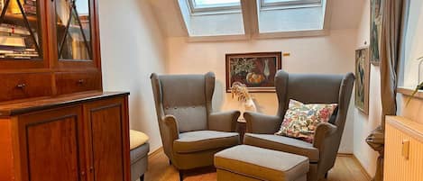 Une chambre à coucher confortable, aménagée en mezzanine, avec un puits de lumière, une armoire en bois, deux fauteuils, une petite table et un radiateur.