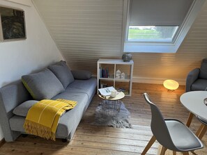 A cozy loft-style room with a gray sofa, a small white side table, a bookshelf, and a skylight.