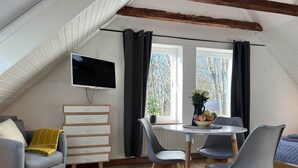 Un dormitori tipus loft acollidor amb un sostre inclinat, un televisor de pantalla plana penjat a la paret, una taula de dinar amb cadires, un prestatge i una finestra amb cortines.