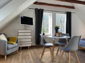 A cozy loft bedroom with a sloped ceiling, a flat-screen TV mounted on the wall, a dining table with chairs, a dresser, and a window with curtains. - Wohnung Atelier - Willi Ohler Haus (Worpswede)
