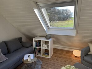 Un dormitorio estilo loft acogedor con techo inclinado, tragaluz, sofá gris, estantería blanca y una mesa redonda de madera.