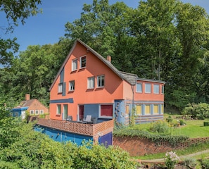 Una casa de dos pisos con fachada roja y azul, rodeada de abundante vegetación y un jardín cuidado.