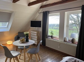 Ein gemütliches Zimmer mit schrägen Wänden, einem runden Tisch mit Stühlen, einem Fernseher und einem Fenster mit Blick auf Bäume.