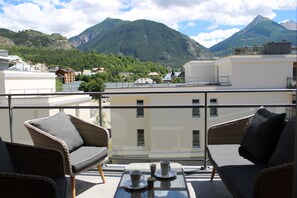 Un balcon agrémenté de meubles en osier et d’une table, offrant une vue sur un paysage montagneux avec des bâtiments en arrière-plan.
