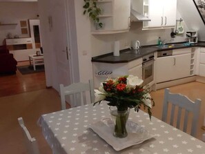 A dining area with a table set for two, a vase of flowers, and a kitchen in the background.
