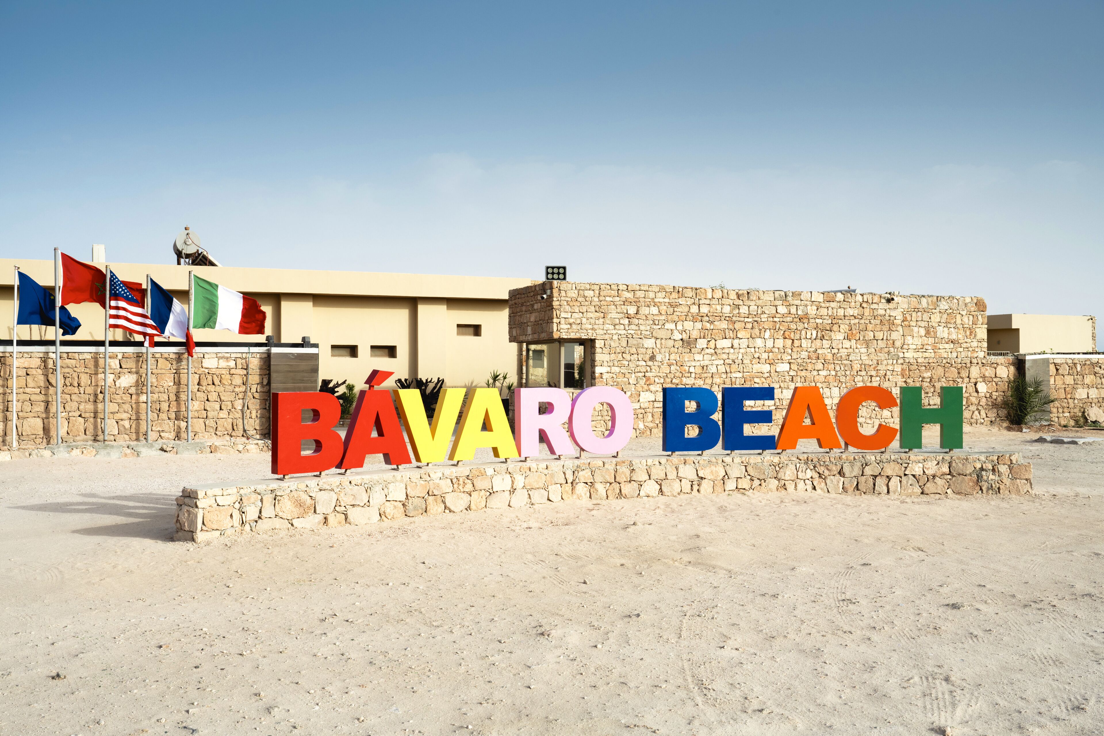 Un cartel de Bávaro Beach con varias banderas y un edificio de piedra al fondo.