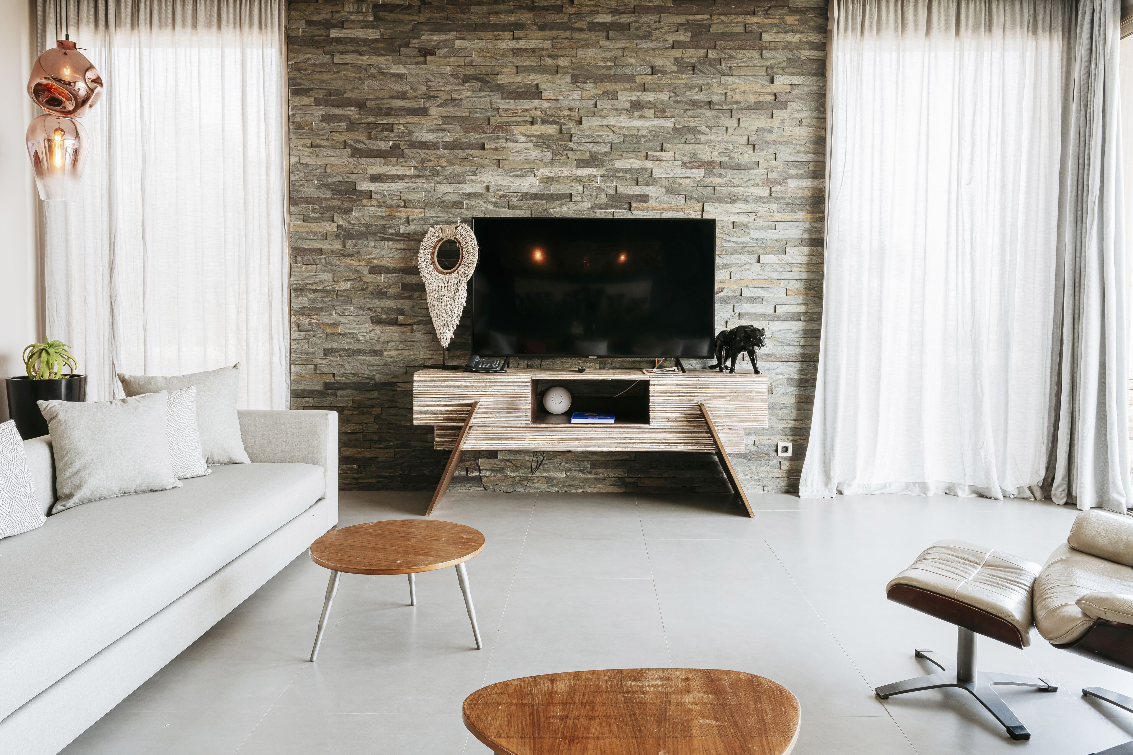 Uma sala de estar moderna com parede de pedra, televisão de tela plana, sofá, mesa de centro e um banquinho de couro.