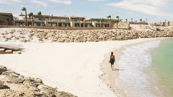A sandy beach with a person walking along the shore, a boat on the sand, and a row of buildings in the background.