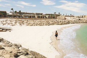 A sandy beach with a person walking along the shore, a boat on the sand, and a row of buildings in the background.