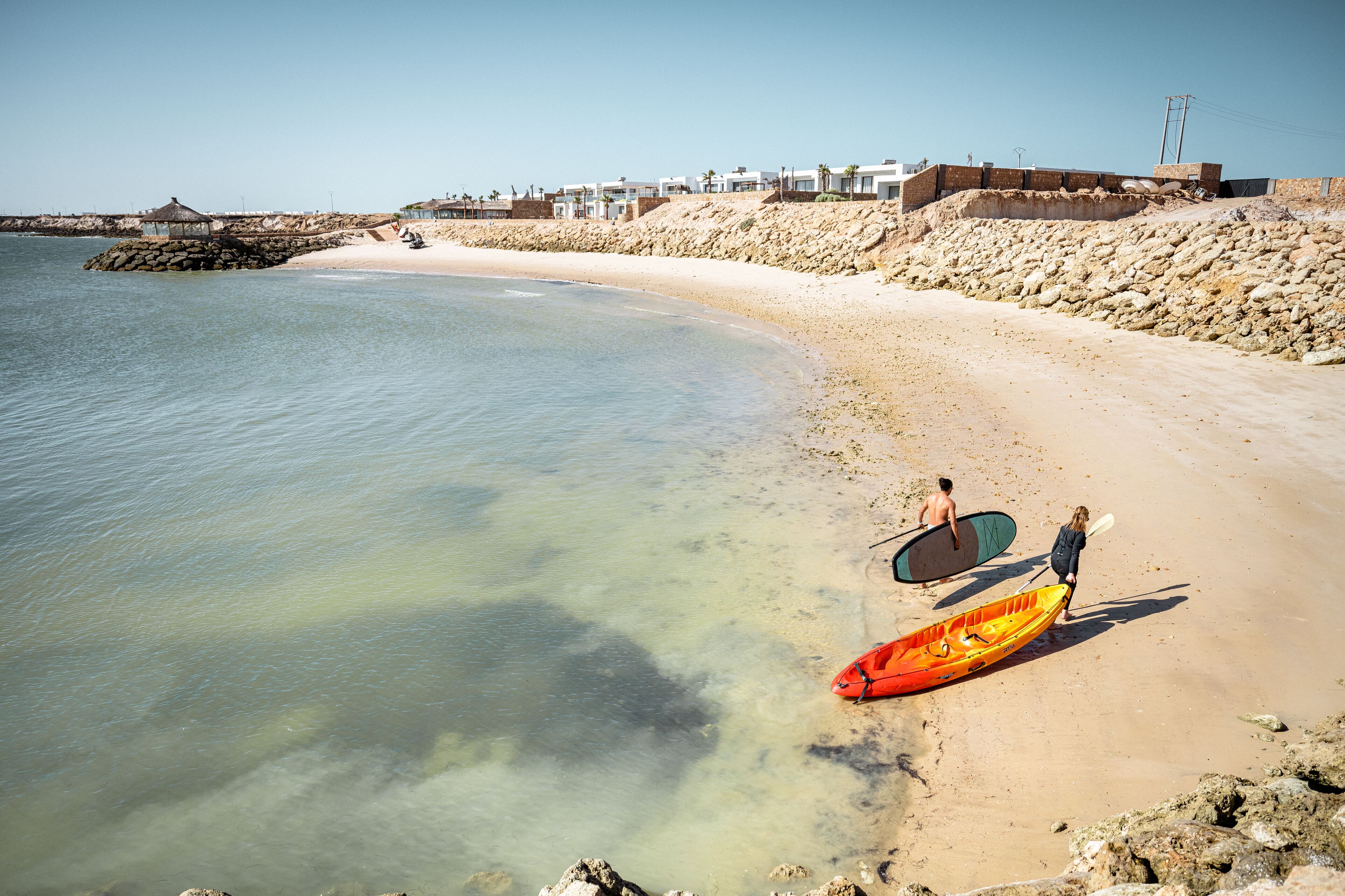 Una playa con agua cristalina, costa rocosa y edificios al fondo.