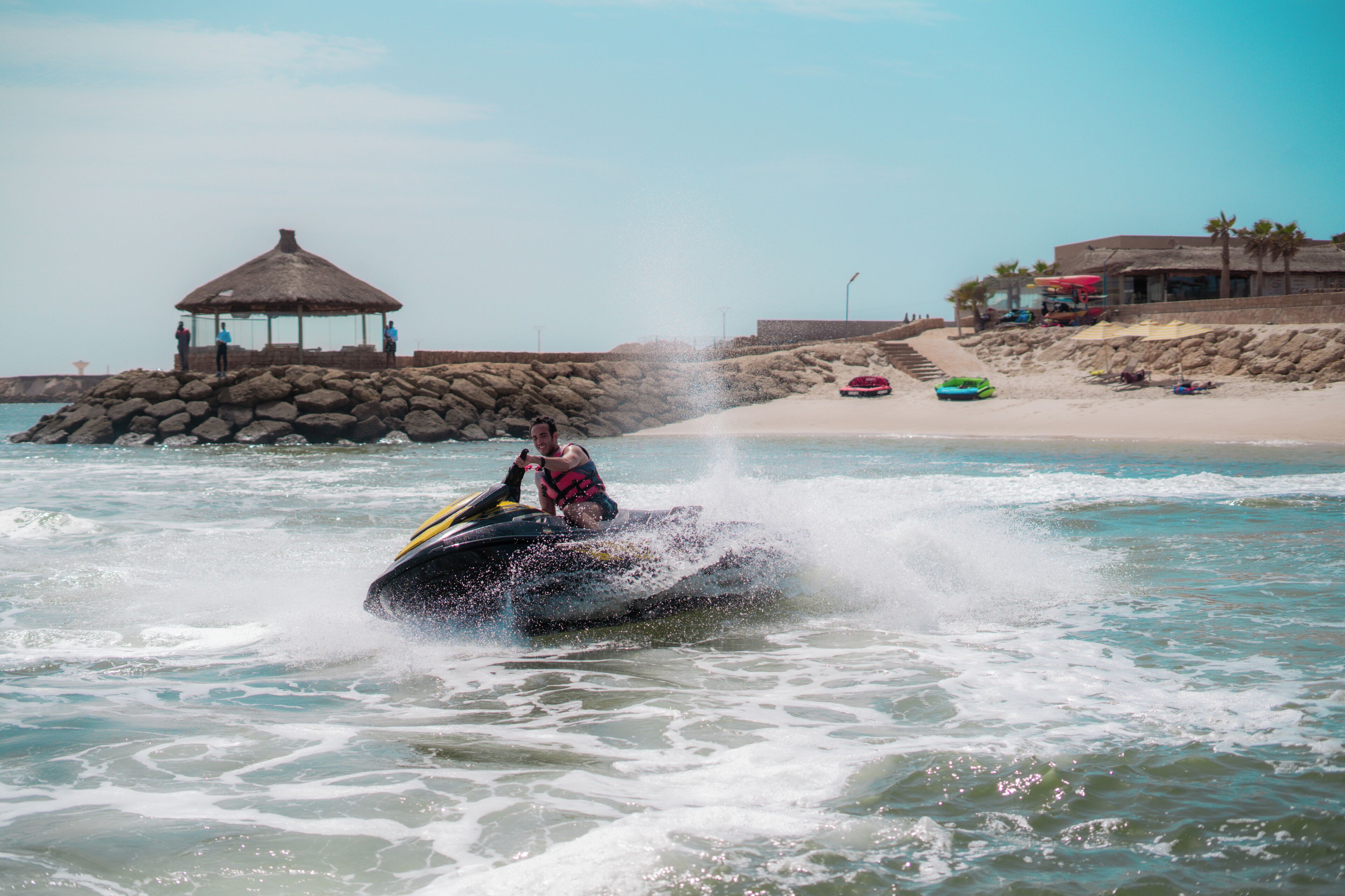 A jet ski rider in the water with a thatched-roof structure and a beach in the background.
