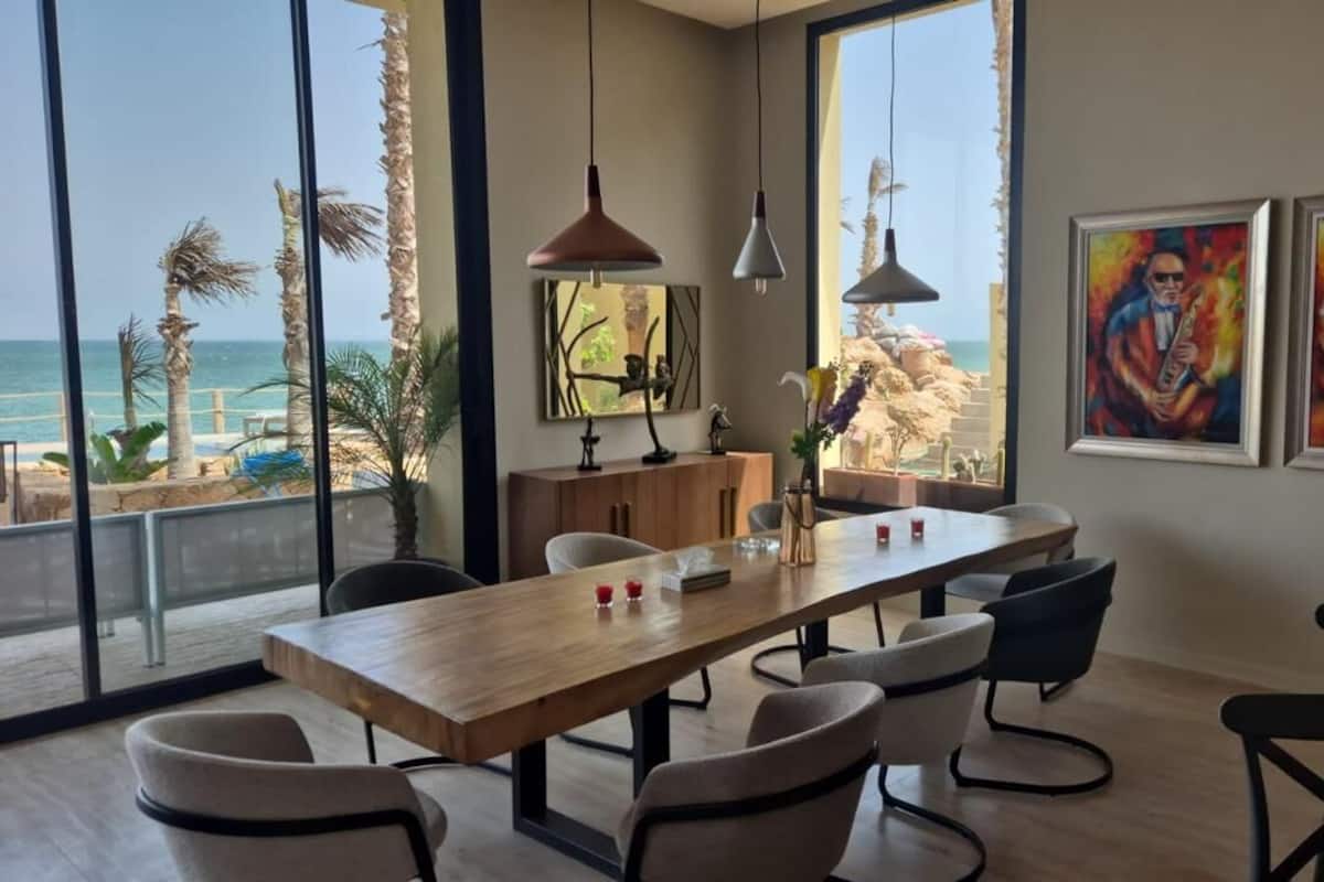 Un espace repas moderne avec une table en bois, des chaises, des luminaires suspendus et de grandes fenêtres donnant sur une plage.