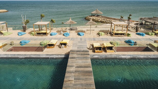 Un espace piscine aménagé au bord de la plage, avec des chaises longues et des parasols, une terrasse en bois et une vue sur la mer.