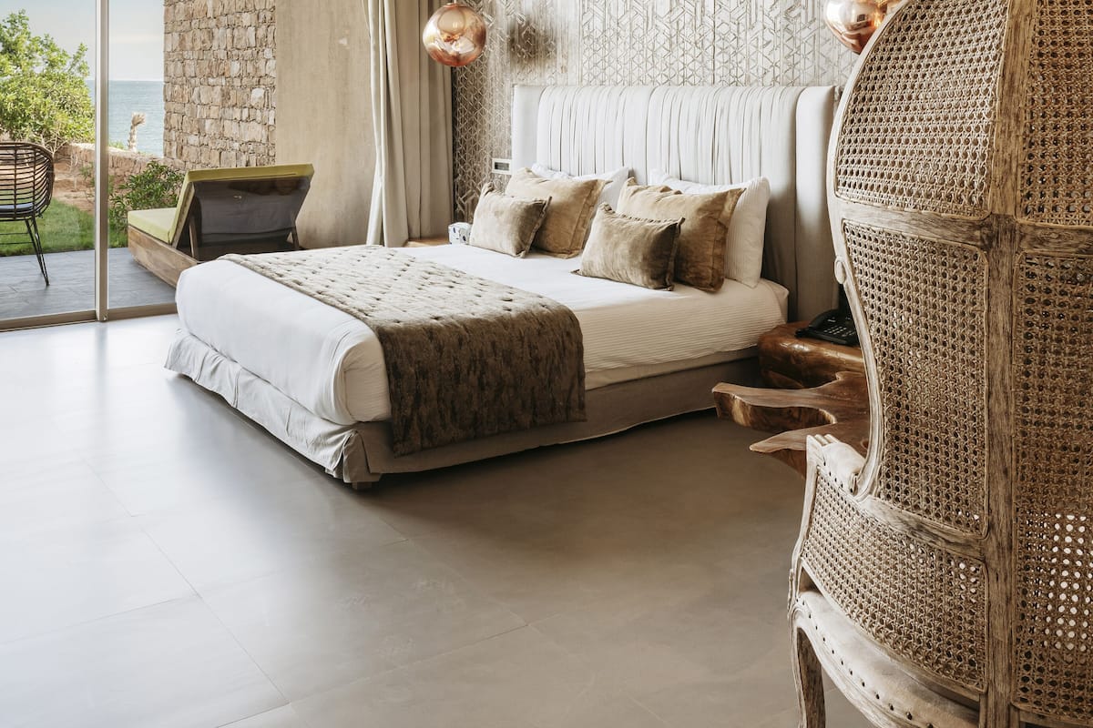 Une chambre moderne avec un grand lit, un fauteuil en rotin, un mur en pierre et une vue sur la mer.
