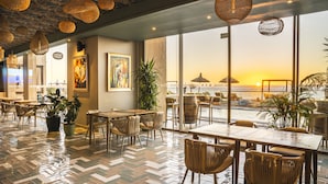 Un restaurant doté d’un sol à motifs en chevrons, de meubles en osier et de grandes fenêtres offrant une vue sur le coucher de soleil et la plage.