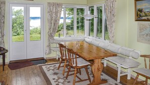 Un espace repas comprenant une table et des chaises en bois, un banc blanc avec coussin, et de grandes fenêtres donnant sur un paysage verdoyant et un plan d’eau.