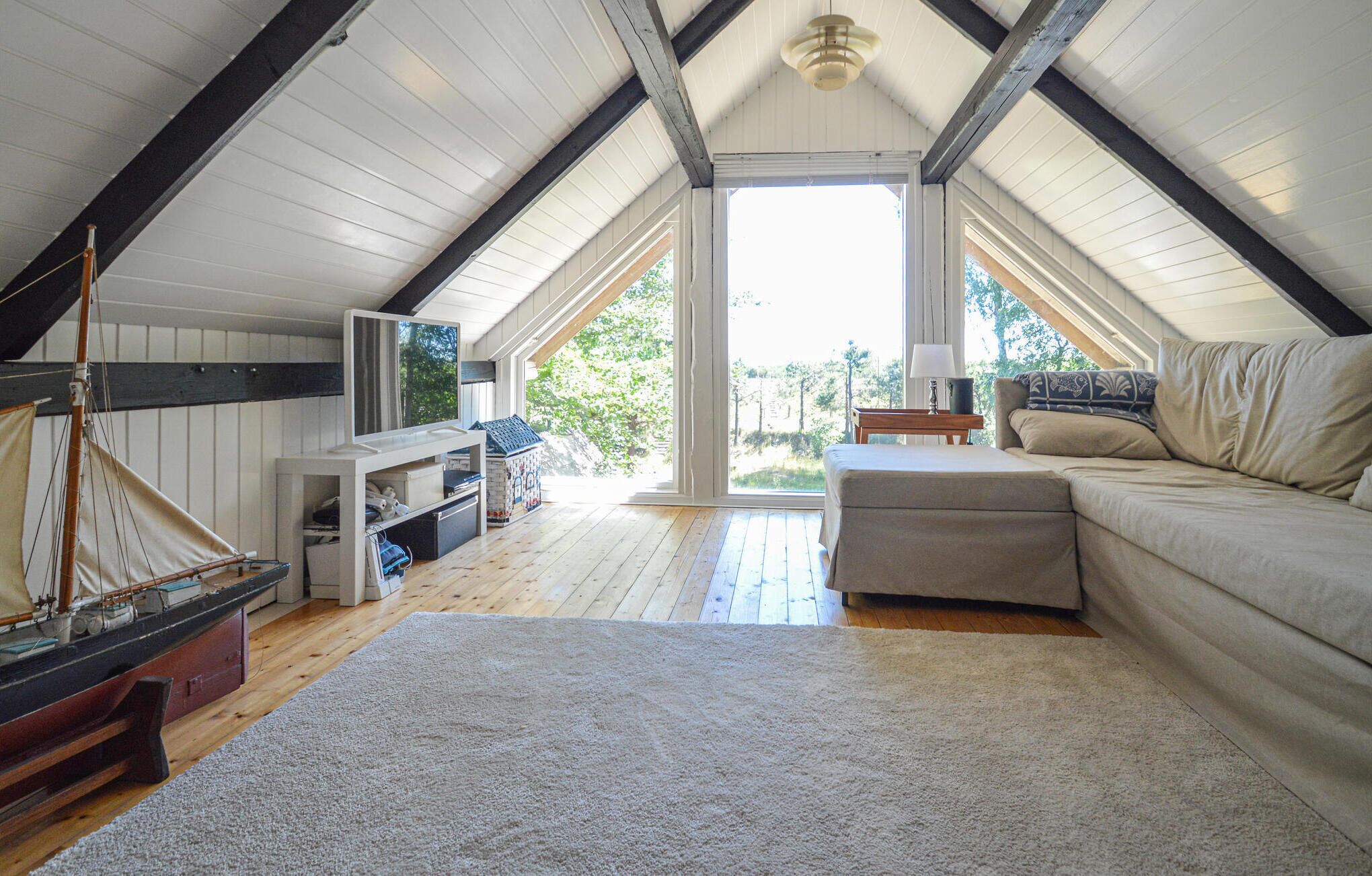 Une chambre mansardée chaleureuse avec un plafond en bois, une grande fenêtre, une maquette de voilier, un canapé et une petite table.