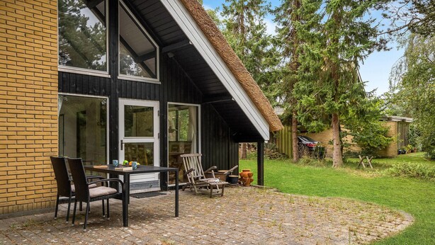 A house with a patio area featuring a table and chairs, a brick wall, and a garden with trees and a shed. - Lovely home in Væggerløse with kitchen (Væggerløse)